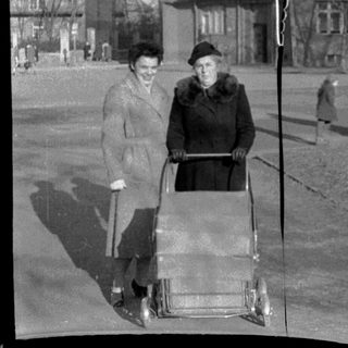 Two women pushing a baby stroller outdoors in urban setting.
