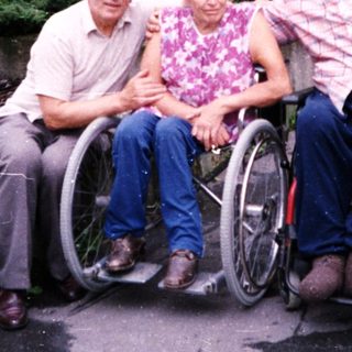 Three elderly individuals in wheelchairs posing outdoors.