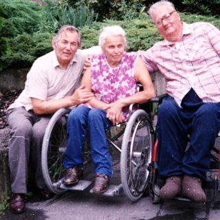 Three elderly individuals in wheelchairs posing outdoors.