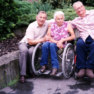 Three elderly individuals in wheelchairs posing outdoors.