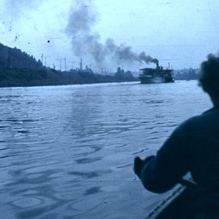 Steamship emitting smoke on a river near industrial shoreline.