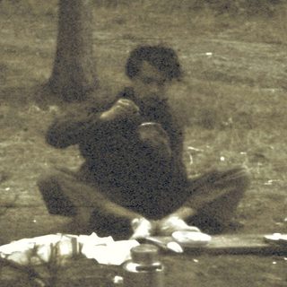 Man sitting outdoors at night, eating near camping supplies.