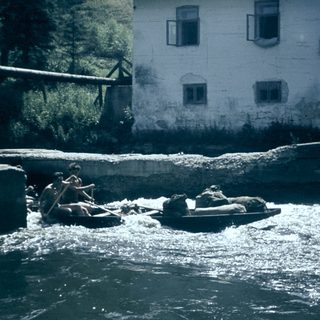 People rafting on turbulent river near old building.