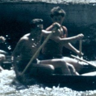 People rafting on turbulent river near old building.