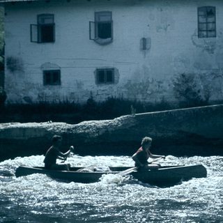 Two people rafting on turbulent river near old building.