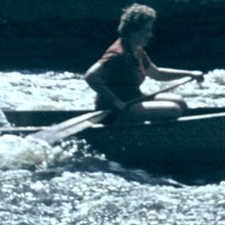 Two people rafting on turbulent river near old building.