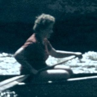 Two people rafting on turbulent river near old building.