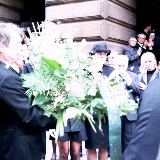 Formal gathering with floral tributes outside building.