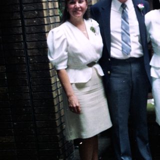 Group of seven people in 1970s-80s attire posing outdoors.
