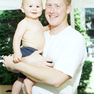 A man holding a smiling toddler outdoors.