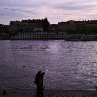Two people by river at dusk near city.