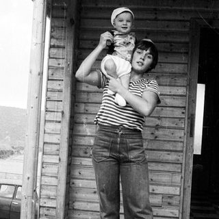 A woman holding a baby outside a wooden shed.
