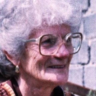 Elderly woman sitting outdoors in patterned dress.