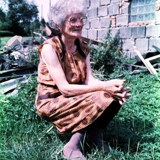 Elderly woman sitting outdoors in patterned dress.