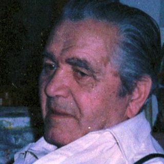 Man sitting indoors wearing suspenders and a button-up shirt.