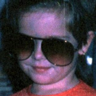 Young child wearing sunglasses, red sweater, and skirt.