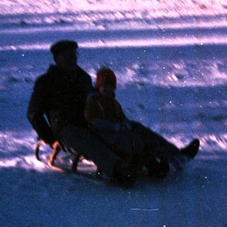 Two people sledding down snow-covered hill at dusk.