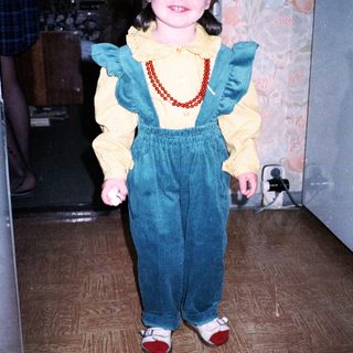 Young girl wearing yellow sweater, blue overalls, and red shoes.
