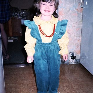 Young girl wearing yellow sweater, blue overalls, and red shoes.