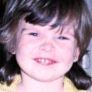 Young girl wearing yellow sweater, blue overalls, and red shoes.