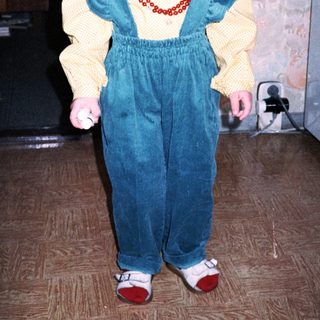 Young girl wearing yellow sweater, blue overalls, and red shoes.