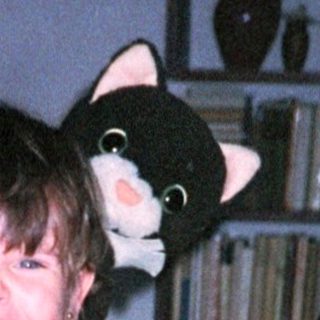Child wearing oversized mittens and overalls, holding stuffed cat.