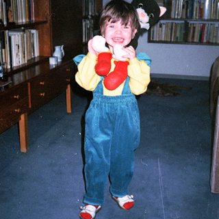 Child wearing oversized mittens and overalls, holding stuffed cat.