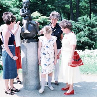 Four women posing with child near Little Mermaid statue.
