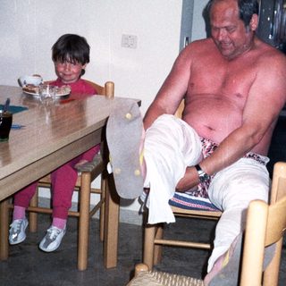 Shirtless man sitting on chair with child nearby.