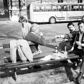 Three men sitting on bench drinking outdoors near buses.