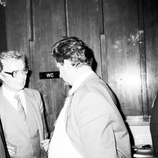 Three men in formal attire conversing indoors near restrooms.