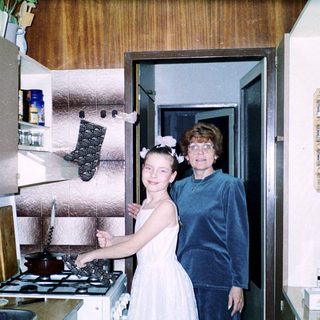 Two women in kitchen: young girl in bridesmaid dress with older woman.