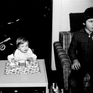 Young child sitting at table eating, older child observing.