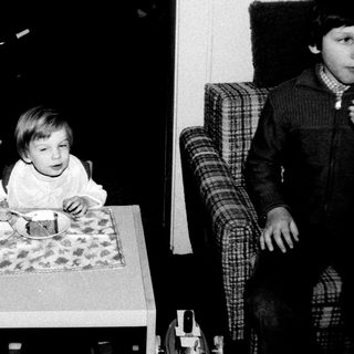 Young child sitting at table eating, older child observing.