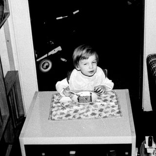 Young child sitting at table eating, older child observing.