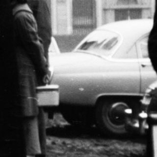Group of people posing outside a building in 1950s-era street scene.
