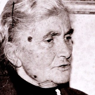 Elderly woman in formal attire, seated indoors.