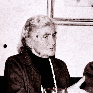 Elderly woman in formal attire, seated indoors.