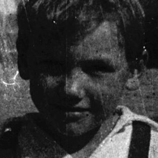 Young boy sitting indoors, wearing suspenders.