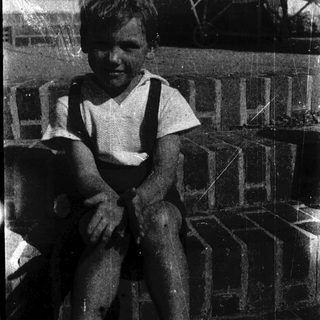 Young boy sitting indoors, wearing suspenders.