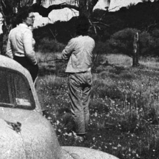 Classic car with vintage license plate, two men working on hood outdoors.
