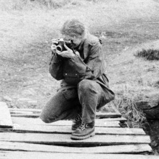 Soldier sitting on makeshift bridge in muddy, war-torn terrain.