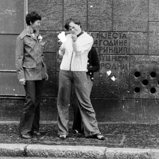 Two men standing outside near a wall with text. One drinks from a cup.