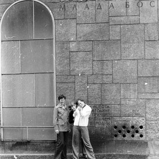 Two men posing with camera near monument plaque in urban setting.