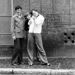 Two men posing with camera near monument plaque in urban setting.
