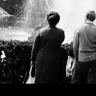 People observing large fountain in grand park setting.