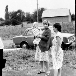 Three women standing outdoors with a baby, holding groceries near vintage cars.