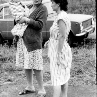 Three women standing outdoors with a baby, holding groceries near vintage cars.