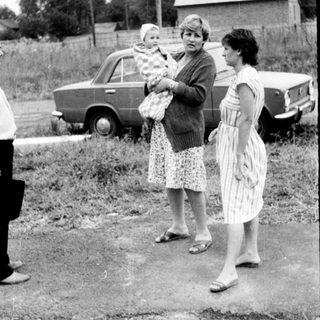 Three women standing outdoors with a baby, holding groceries near vintage cars.