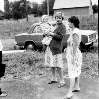 Three women standing outdoors with a baby, holding groceries near vintage cars.